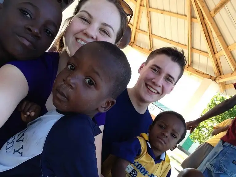 Five people, including two adults and three children, smile and pose for a group selfie under a wooden pavilion as one of the kids waves enthusiastically at the camera.