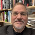 Matthew Becker, Ph.D., M.Div., a man with glasses and a beard, smiles in front of shelves filled with books and papers.