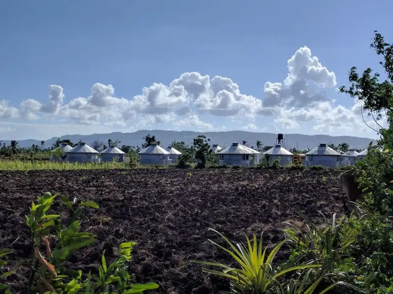 White round houses with silver roofs stand behind a plowed field, under a bright blue sky with clouds shaped like gentle waves.