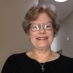 Smiling older woman with glasses, wearing a black top and cross necklace, standing indoors.