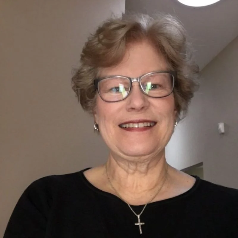 Smiling older woman with glasses, wearing a black top and cross necklace, standing indoors.
