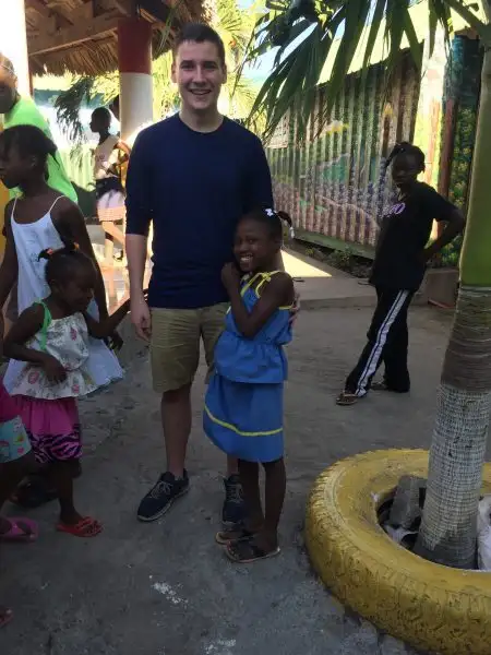 A man and a smiling young girl pose together outdoors, surrounded by other children near a painted tire planter, as the girl waves happily at the camera.