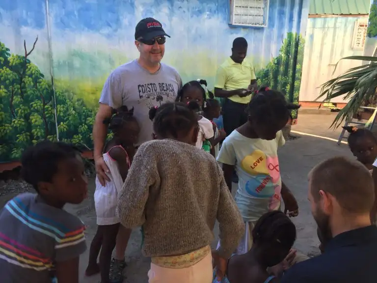 Man in sunglasses and cap stands with a group of smiling children outdoors near a mural and colorful container buildings, all enjoying the positive WAVES of energy in the vibrant setting.
