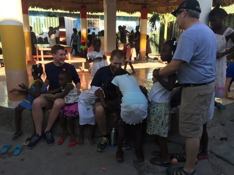 Adults and children interact and play together outdoors in a sunny, shaded area, as laughter and cheerful WAVES of activity fill the background.