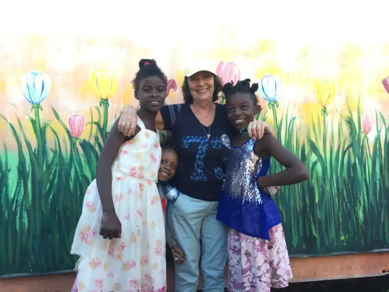 A woman and three smiling children stand in front of a colorful flower mural, posing for a photo as one child WAVES cheerfully at the camera.