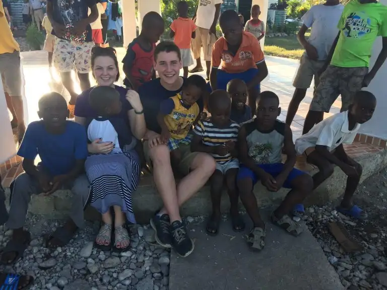 Two adults sit on a step, surrounded by smiling children outdoors on a sunny day, as laughter and WAVES of joy fill the air.