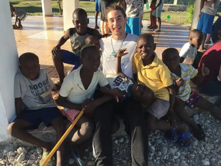 Smiling young man sitting outdoors, surrounded by happy children on a sunny day, as gentle WAVES of laughter fill the air.