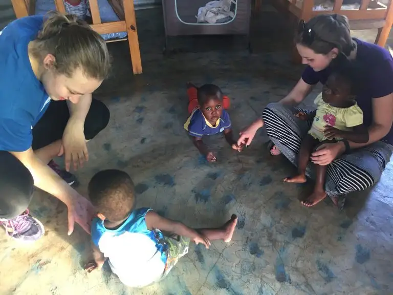 Two women interact with three young children sitting and crawling on a blue patterned WAVES floor indoors.