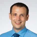 Smiling man in a light blue dress shirt and tie against a plain, light background—Jesse Sestito, Ph.D.