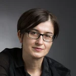 Slavica Jakelić, a woman with short brown hair and glasses, wearing a black shirt, poses against a gray background.