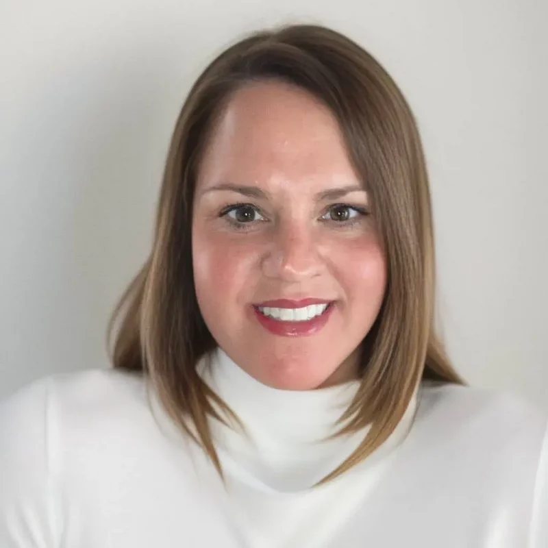 Jamie Stangel, Ph.D., with straight brown hair and a white turtleneck, smiles in front of a plain light background.