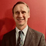Smiling man in a suit and tie, identified as Jeffrey Emmons, standing in front of a red wall.