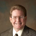 Smiling man with glasses and short hair wearing a suit and tie in front of a brown background, identified as Jeffrey Scott Doebler.