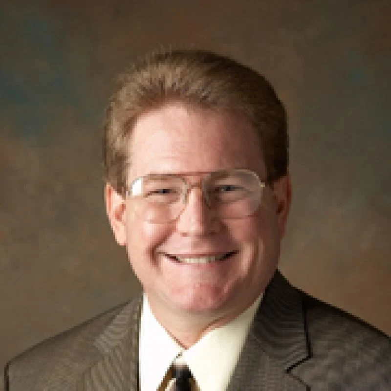 Smiling man with glasses and short hair wearing a suit and tie in front of a brown background, identified as Jeffrey Scott Doebler.