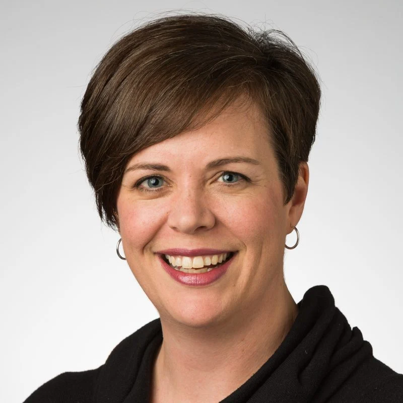 Smiling woman with short brown hair, wearing hoop earrings and a black top, on a light background—Kristin Lewis, M.A.