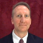 Smiling man in a suit and tie with short hair, posing in front of a red textured background, introduces himself as Karl D. Madsen, MBA.
