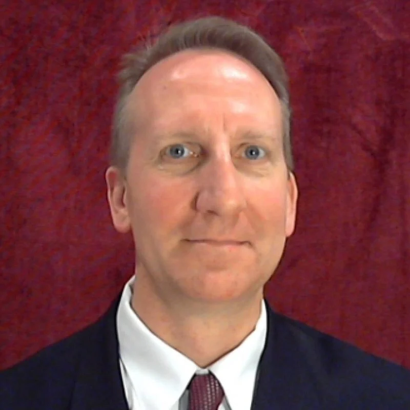 Smiling man in a suit and tie with short hair, posing in front of a red textured background, introduces himself as Karl D. Madsen, MBA.