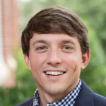 Lucas Kelley, a young man smiling outdoors in a navy blazer and checkered shirt, stands against a backdrop of lush greenery.
