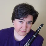 Denise Chigas Kirkland, with short dark hair in a purple shirt, holds a clarinet and looks at the camera.