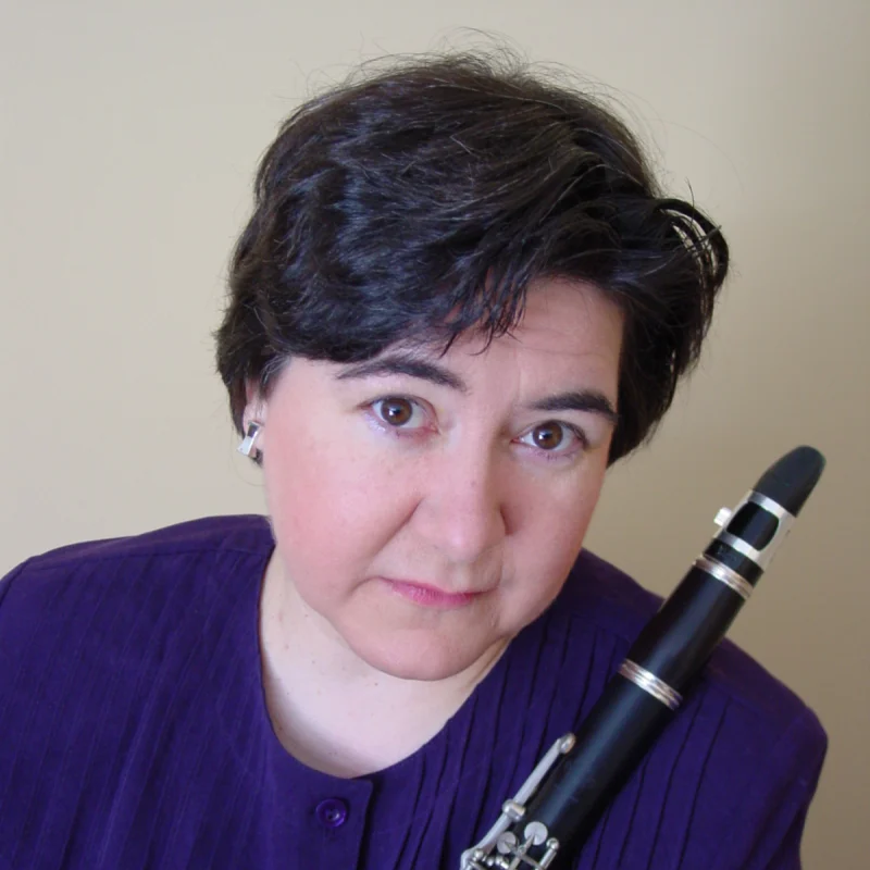 Denise Chigas Kirkland, with short dark hair in a purple shirt, holds a clarinet and looks at the camera.