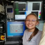 Leanne Blind-Doskocil smiles in front of multiple computer screens displaying technical and weather data in a control room.