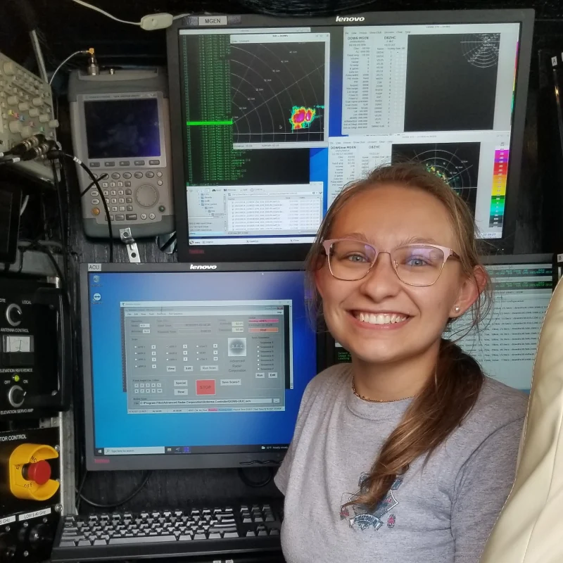 Leanne Blind-Doskocil smiles in front of multiple computer screens displaying technical and weather data in a control room.