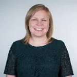 Smiling woman with straight blonde hair, MA, Elizabeth Corcoran, wearing a dark green lace top, standing against a plain light background.