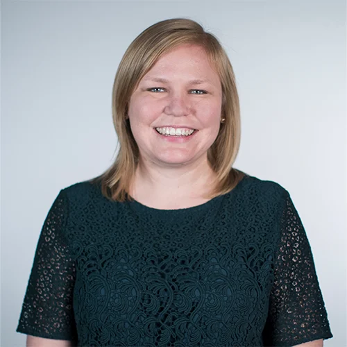 Smiling woman with straight blonde hair, MA, Elizabeth Corcoran, wearing a dark green lace top, standing against a plain light background.