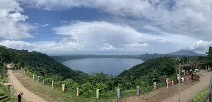 Panoramic view of a blue lake with gentle waves, surrounded by green hills, with people and horses along a path in the foreground.