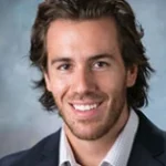 Smiling man with medium-length brown hair in a dark jacket and light shirt, posing against a gray background—David Lantis, PhD.