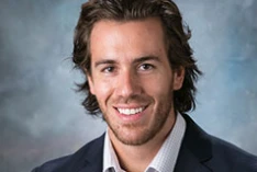 Smiling man with medium-length brown hair in a dark jacket and light shirt, posing against a gray background—David Lantis, PhD.