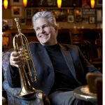 Smiling man in a suit holding a trumpet while sitting in a cozy, warmly lit café or bar.