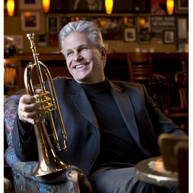 Smiling man in a suit holding a trumpet while sitting in a cozy, warmly lit café or bar.