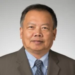 A man in a brown suit and blue shirt poses against a plain light gray background.