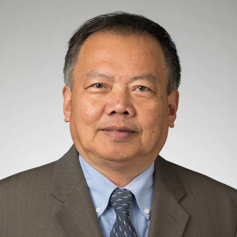 A man in a brown suit and blue shirt poses against a plain light gray background.