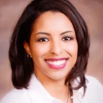 Lacey Logan, Ph.D., with shoulder-length dark hair, smiles warmly in a white shirt, standing in front of a blurred background.