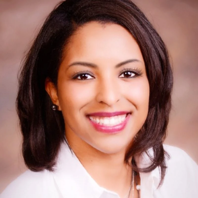 Lacey Logan, Ph.D., with shoulder-length dark hair, smiles warmly in a white shirt, standing in front of a blurred background.