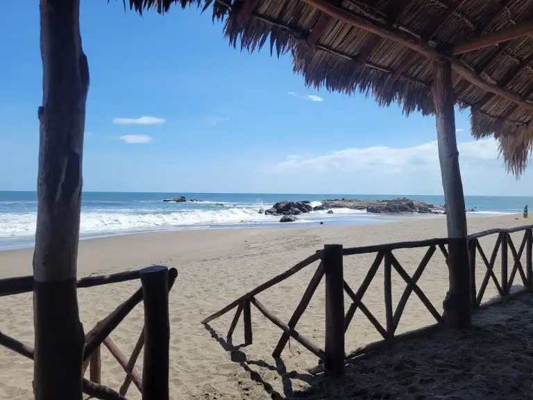 View of gentle waves rolling onto a sandy beach, seen from beneath a thatched-roof hut with a wooden fence.