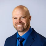 Smiling bald man with a beard, Vince Lusk, M.A., in a blue suit and matching shirt against a light background.