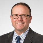 Matthew Luth, Ph.D., wearing glasses, a gray jacket, checked shirt, and blue tie, smiles warmly against a plain background.