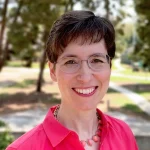 Marin Jacobson, a woman with short brown hair, glasses, and a pink shirt, smiles outdoors with trees in the background.