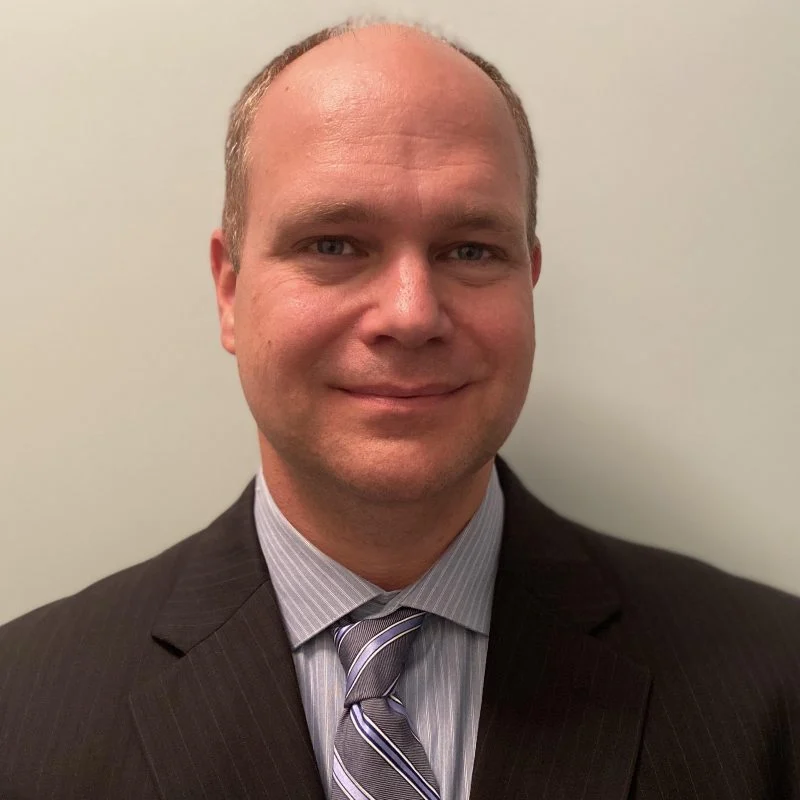 Matt Campbell, dressed in a suit and striped tie, smiles warmly in front of a plain light-colored background.