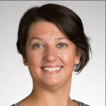 Smiling woman with short brown hair, wearing a patterned top, stands against a plain light background—Natalie Muskin-Press.