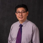 Peixing Jiang, Ed.D., wearing glasses, a light purple shirt, and a patterned tie, poses confidently in front of a dark background.