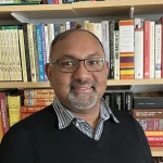 Smiling man with glasses and beard, likely George Pati, Ph.D., standing in front of a bookshelf filled with colorful books.