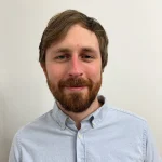 Paul Olander, a man with light brown hair and a beard, wears a light blue button-up shirt while standing against a white wall.