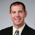 Smiling man in a suit and tie with short hair, posing against a plain light gray background—introducing Paul Rowe, Ph.D.