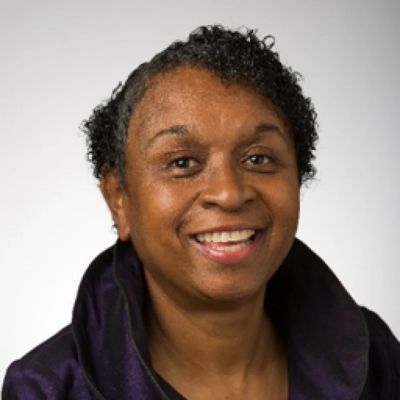 Smiling woman with short curly hair wearing a dark purple collared top, posing against a light background.
