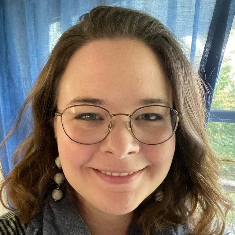 Woman with glasses and wavy brown hair smiling in front of a window with blue curtains.