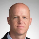 A bald man in a suit and collared shirt, resembling Matthew Puffer, looks at the camera with a neutral expression.